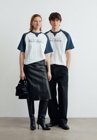 Γυναίκα και άνδρας στέκονται, φορώντας ταιριαστά λευκά και ναυτικά ριγέ πουκάμισα· η γυναίκα φορά μαύρη δερμάτινη φούστα και μπότες και κρατά μαύρη τσάντα χειρός.