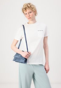 White cotton t-shirt with a chest pocket, paired with light blue trousers and a small blue suede handbag with a double-flap design.