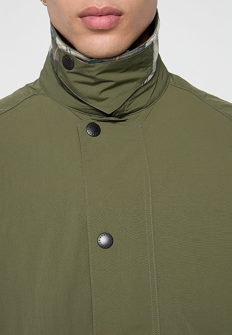 Olijfgroene jas met een hoge kraag, zwarte drukknoopjes en een patroon in de binnensluiting van de kraag. Heeft een gestructureerde stof en zichtbare stiksels.