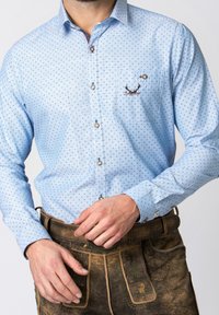 Muž v světle modré vzorované košili s knoflíky a vyšitými parohy jelena na kapse, zasunuté do tradičních hnědých kožených kalhot.