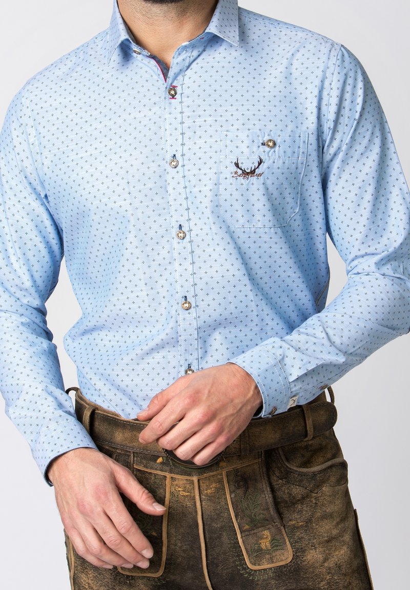 Muž v světle modré vzorované košili s knoflíky a vyšitými parohy jelena na kapse, zasunuté do tradičních hnědých kožených kalhot.