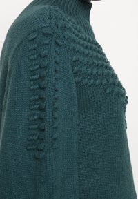 Pull en maille bleu sarcelle foncé avec col montant. Comprend des accents texturés le long des épaules et des côtés. Confectionné en mélange doux de laine.