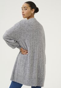 Graue Strickjacke mit geripptem Saum, versehen mit einem strukturierten Kabelmuster und weiten Ärmeln, die bis zur Mitte des Oberschenkels reichen.