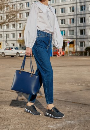 Personne marchant sur le trottoir portant un chemisier blanc et un jean bleu, portant un grand sac à main bleu marine, avec des voitures et un bâtiment en arrière-plan.