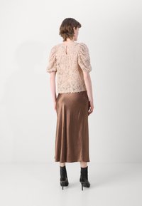 Spitzenoberteil in hellem Beige mit aufgesetzten Ärmeln, kombiniert mit einem braunen Satinrock. Das Model trägt schwarze, heeled Stiefeletten. Rückansicht.