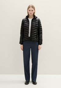 Chaqueta de plumas negra con textura acolchada, diseño ajustado y capucha. Combinada con pantalones de pierna ancha azul oscuro y calzado negro. Fondo neutro.