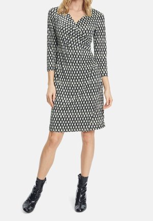 Mujer con un vestido a la rodilla de patrón geométrico en blanco y negro, mangas tres cuartos y botines de charol negro.