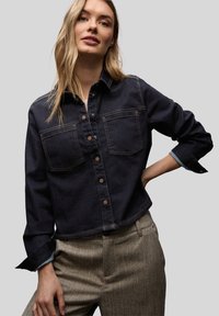 Femme portant une chemise en jean foncé à boutons avec poches avant et un pantalon beige texturé taille haute, posant avec la main sur la hanche.