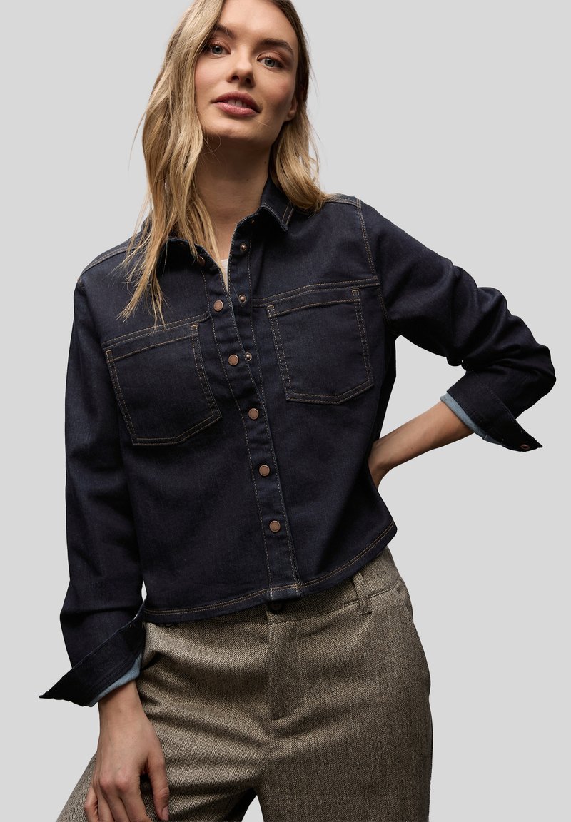 Femme portant une chemise en jean foncé à boutons avec poches avant et un pantalon beige texturé taille haute, posant avec la main sur la hanche.