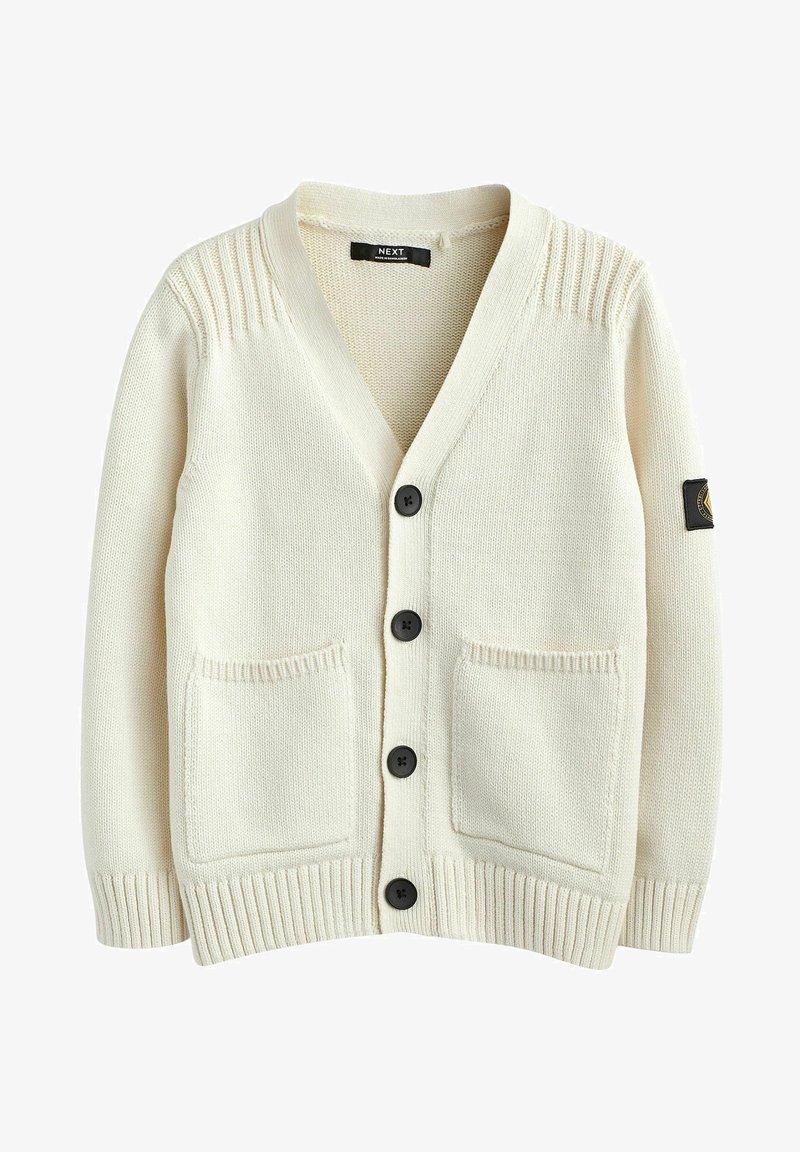 Cardigan en maille de couleur crème avec quatre boutons noirs, deux poches devant, des poignets côtelés et un patch noir sur la manche gauche.