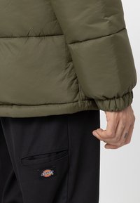 Kurtka puchowa w oliwkowej zieleni o pikowanej fakturze i elastycznych mankietach, noszona z czarnymi spodniami z naszywaną kieszenią i detalem z logo.