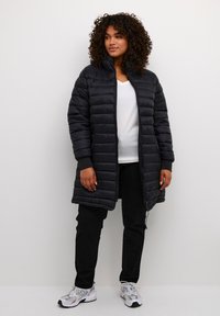 Frau mit lockigem Haar, die eine schwarze gesteppte lange Jacke, ein weißes Oberteil, schwarze Hosen und weiße Sneakers trägt, steht vor einem schlichten Hintergrund.