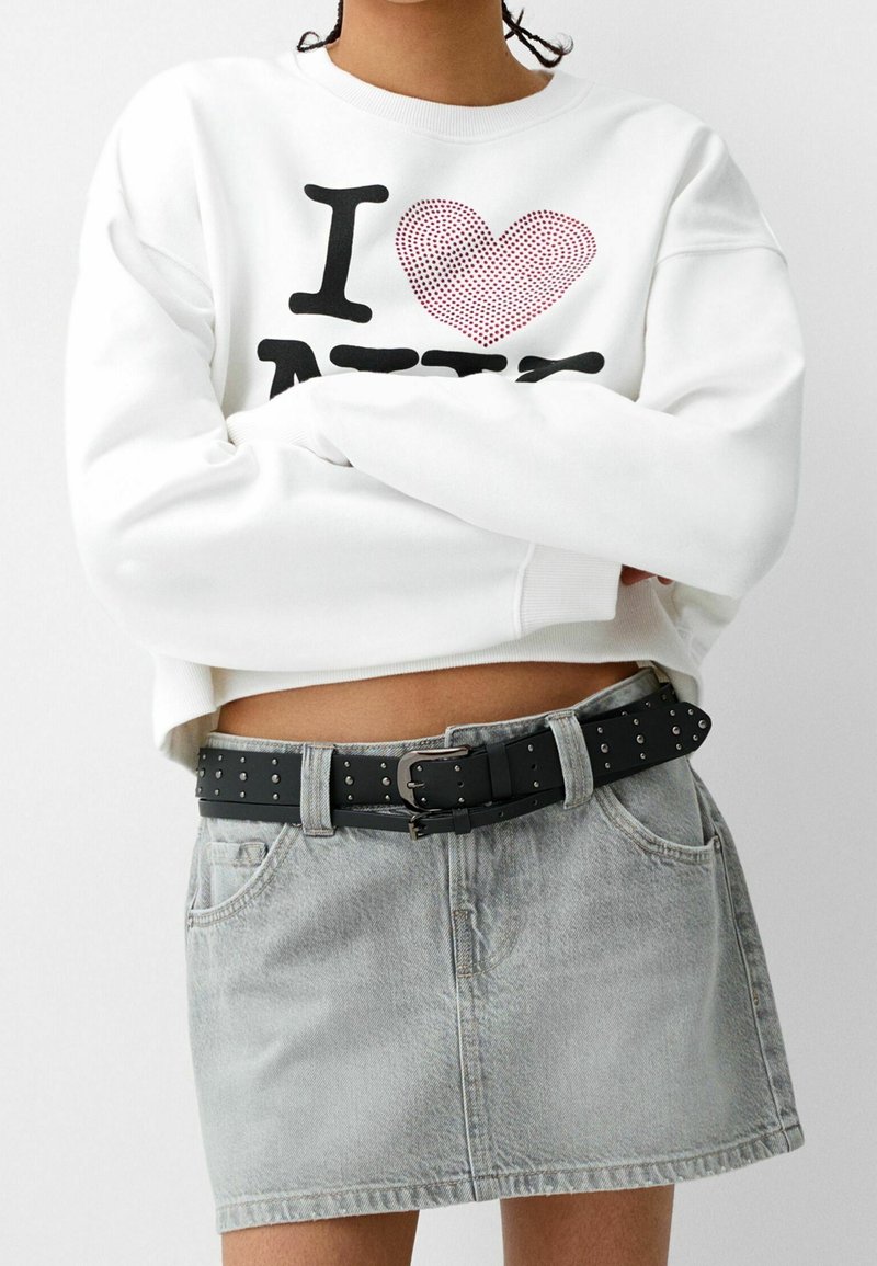 Personne portant un sweat court blanc avec le texte "I heart NY", une jupe en jean grise et une ceinture noire cloutée avec une double boucle.