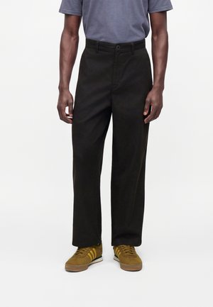 Homme portant un pantalon noir droit, un t-shirt gris et des baskets jaune moutarde avec des rayures blanches et vertes, debout sur un fond blanc.