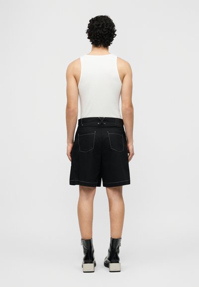 Άνδρας με σγουρά σκούρα μαλλιά, στέκεται γυρισμένος με την πλάτη, φορώντας λευκό αμάνικο μπλουζάκι, μαύρα σορτς με εμφανείς ραφές και μαύρα μποτάκια μέχρι τον αστράγαλο.