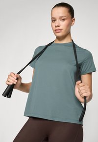 Jovem mulher vestindo uma camisa azul-turquesa e leggings castanhas segura uma corda de saltar preta sobre os ombros, contra um fundo simples.