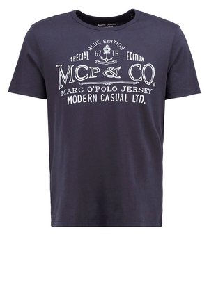Mann trägt blaue gesteppte Jacke, schwarzes Grafik-T-Shirt, beige Hose und braune Schnürschuhe, steht mit einer Hand in der Tasche.