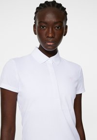 Vit polo-shirt med texturerad tyg, korta ärmar och krage. Har en liten rosa logga på bröstet.