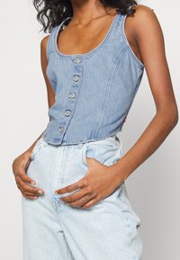 Ljusblå denim ärmlös topp med skjortknappar framtill, figurnära design och en rundad halsringning. Kombinerad med ljusblå denim jeans.