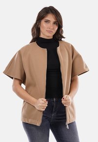 Giacca corta beige con maniche corte e cerniera frontale. Tessuto uniforme, materiale liscio e design minimalista. Indossata sopra una maglietta nera.
