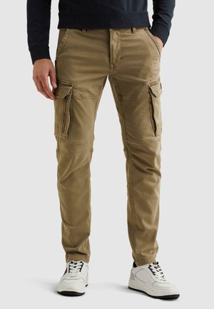 Homme portant un pantalon cargo kaki avec plusieurs poches, les mains dans les poches, assorti de baskets blanches, debout devant un fond uni.