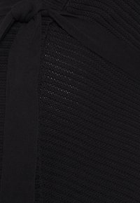 Svart stickat tyg med ett strukturerat mönster, som har en knuten banddetalj. Materialet ser mjukt ut med en svag lyster.