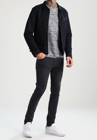 Schwarze Jeansjacke über einem grauen strukturierten Pullover, kombiniert mit dunklen Jeans und weißen Sneakern, getragen von einer Person, die lässig steht.