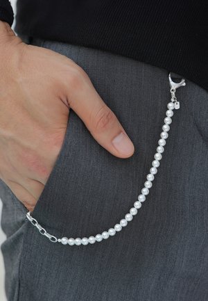 Sølvkæde med perlelignende perler, fastgjort til en lomme. En hånd er delvist synlig og hviler mod gråt stof. Simpel låse detalje.