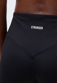 Κοντινό πλάνο της κάτω πλάτης και μέσης ενός ατόμου που φοράει μαύρα κολάν με τη λέξη "STRONGER" τυπωμένη σε λευκό στη μέση.