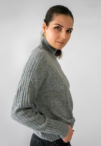 Grijze trui met een hoge geribbelde kraag, gestructureerde mouwen en een relaxte pasvorm, gecombineerd met donker jeans, tegen een neutrale achtergrond.