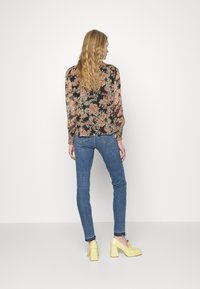 Blusa floral com mangas longas e bufantes em preto, laranja e azul-turquesa; combinada com jeans justos azuis e sapatos de salto bloco amarelos.