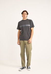 Camiseta de algodón gris oscuro con un patrón decorativo horizontal en gris claro, combinada con pantalones oscuros de corte holgado en verde olivo y zapatillas negras.