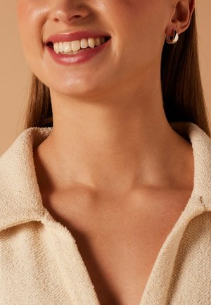 Vrouw die glimlacht en tanden laat zien, draagt kleine oorringen en een beige, textuurrijke kraagdrager.