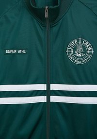 Dark green zip-up jacket with white stripes, featuring an embroidered logo and "UNFAIR ATHL." patch on the left side. Textured fabric.