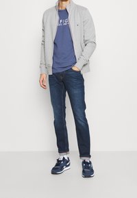 Hellgraue Zip-Jacke mit hohem Kragen, marineblaues T-Shirt mit weißem Text, dunkelblaue Jeans und marineblaue Sneakers mit weißen Akzenten.