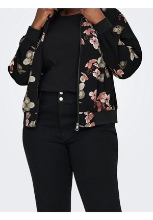 Person iført en sort blomsterbroderet lynlåstrøje over en sort ribbet skjorte og sort højtaljede bukser med to synlige knapper.