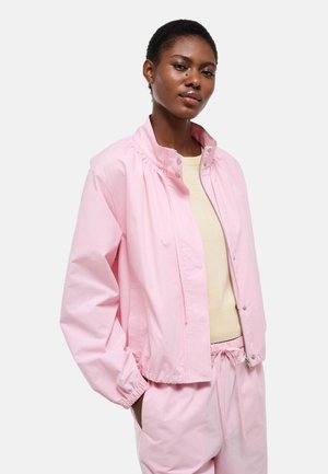 Jeune femme noire portant une veste rose pâle et un pantalon assorti avec un haut beige, debout les mains dans les poches devant un fond uni.