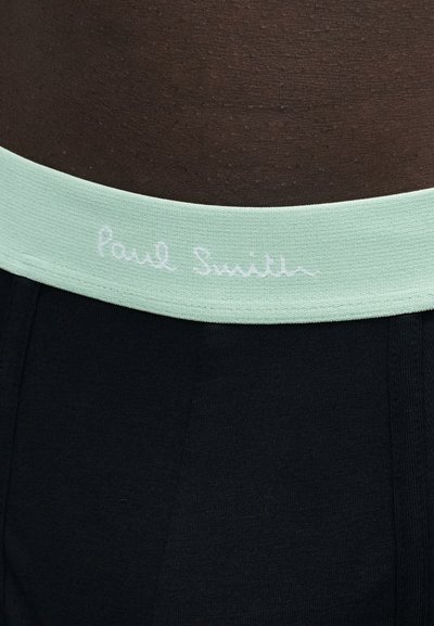 Gros plan sur une personne portant un pantalon noir avec une ceinture vert clair comportant le logo manuscrit blanc "Paul Smith."