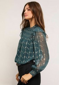 Türkiz áttetsző blúz fémes virágmintával, fodros gallérral és gyűrt mandzsettával. Laza fazonú, hosszú ujjú és bonyolult ráncolt részletekkel rendelkezik.