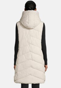 Femme de dos, portant un long gilet matelassé beige sans manches avec une grande capuche, et un haut à manches longues noir en dessous.