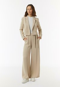 Beige dubbelzijdig jasje met schoudervullingen, gecombineerd met hooggeplaatste, wijde broek in dezelfde kleur en witte platform sneakers.