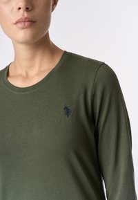 Camicia a maniche lunghe di colore verde oliva in tessuto soffice con scollatura rotonda. Presenta un piccolo logo ricamato sul petto.