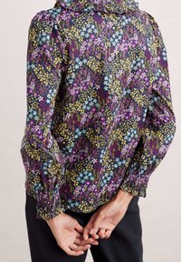 Blomstret bluse med flæset krave, der har flerfarvede blomster på en mørk baggrund. Lange ærmer med elastiske manchetter, tætsiddende pasform.