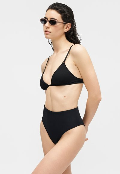 Γυναίκα με βρεγμένα μαλλιά που φοράει μαύρο μαγιό με ψηλόμεσο κάτω μέρος, μαύρο τρίγωνο τοπ μαγιό και καφέ γυαλιά ηλίου σε απλό ανοιχτό φόντο.