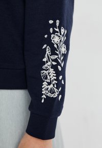 Sudadera de color navy con bordado floral blanco en la manga, que cuenta con un puño acanalado y un tejido suave y texturizado.