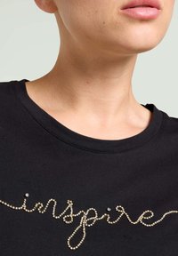 Camiseta de algodón negra con cuello redondo, que presenta la palabra "inspire" en adornos de strass en tono dorado. Textura suave, diseño casual.