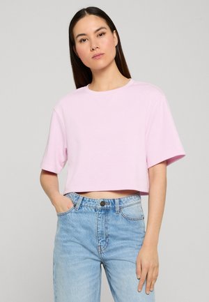 Femme aux longs cheveux foncés portant un t-shirt court rose clair et un jean taille haute bleu clair, debout avec une main dans la poche.