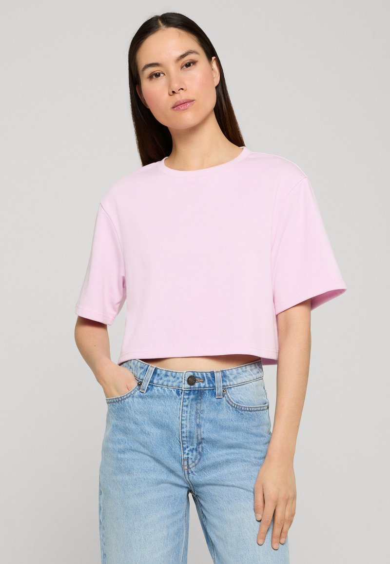 Femme aux longs cheveux foncés portant un t-shirt court rose clair et un jean taille haute bleu clair, debout avec une main dans la poche.