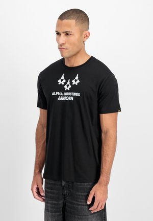 Mann mit kurzen Haaren und Ohrring, trägt ein schwarzes Alpha Industries Airborn T-Shirt und dunkle Jeans, steht vor einem einfarbigen Hintergrund.