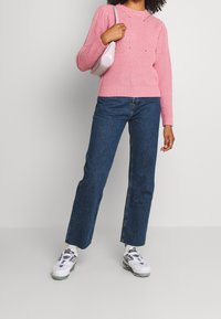 Pull rose en tricot avec texture côtelée, associé à un jean bleu foncé à jambes droites et des baskets blanches. Accessoires comprenant un petit sac à main rose pâle.